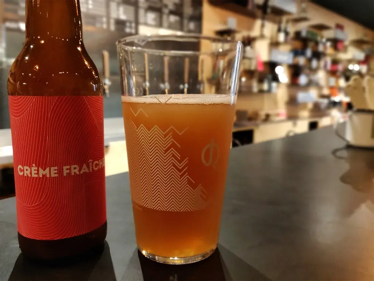 Bière artisanale "Crème Fraîche" dans un verre, avec une bouteille à côté. L'arôme subtil du houblon se marie à la douceur crémeuse.