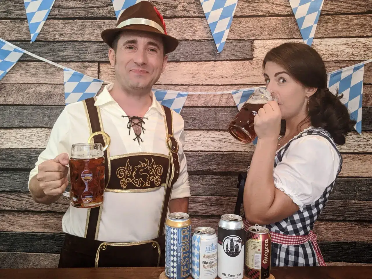 Un homme et une femme en tenue bavaroise c&eacute;l&egrave;brent avec des bi&egrave;res allemandes. Des fanions bleus et blancs d&eacute;corent le fond en bois.