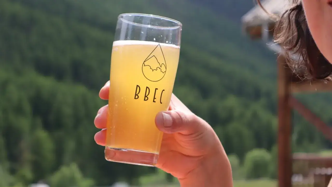 Verre de bi&egrave;re blonde p&eacute;tillante, logo BBEC, avec un paysage de montagne verdoyant en arri&egrave;re-plan. Parfait pour une bi&egrave;re printemps.