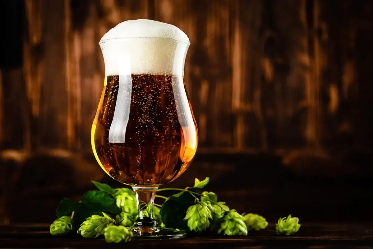 Verre de bière IPA débordant de mousse, avec des cônes de houblon frais sur un fond en bois sombre.