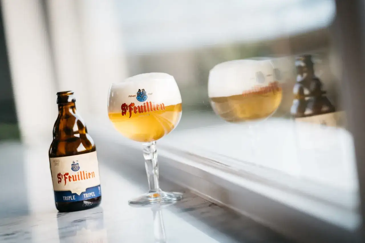 Une bouteille et un verre de bière belge St. Feuillien Triple, avec une mousse généreuse, reflétés sur une surface.