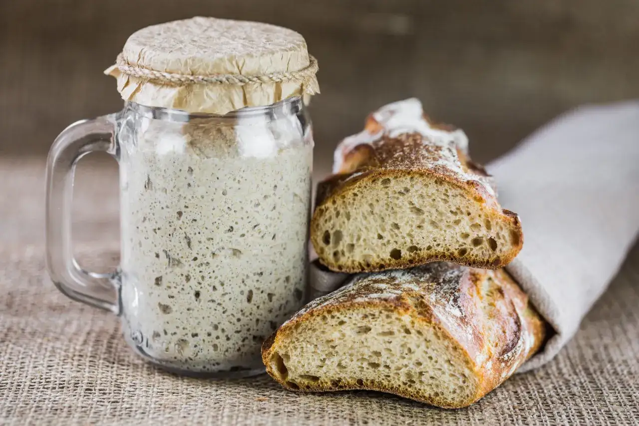 Un bocal de levain pétillant à côté de pains artisanaux coupés. L'art de remplacer la levure par levain pour un goût authentique.