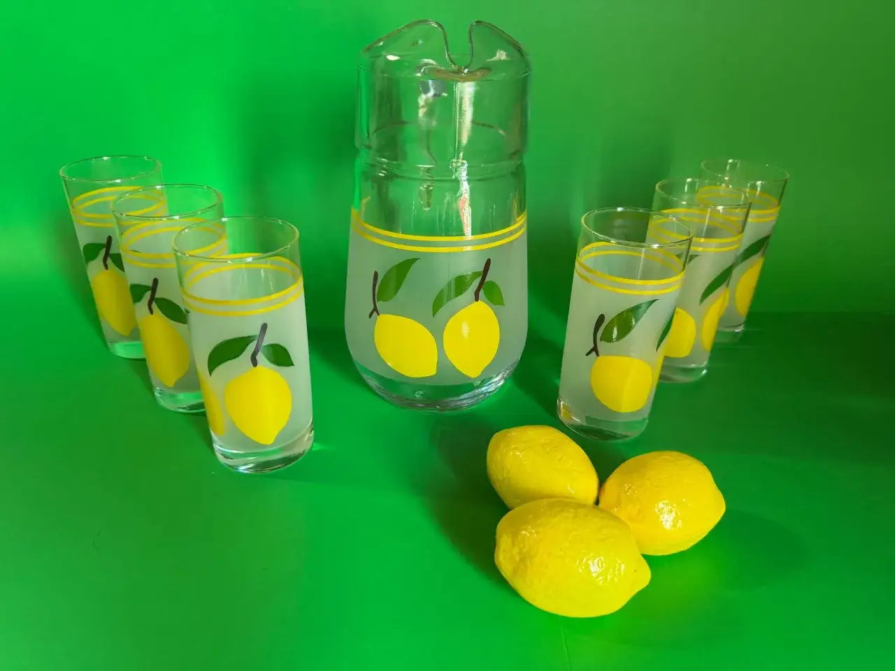 Service à bière citron nom, avec un pichet et six verres décorés de citrons jaunes et de feuilles vertes. Trois citrons frais sont posés devant.