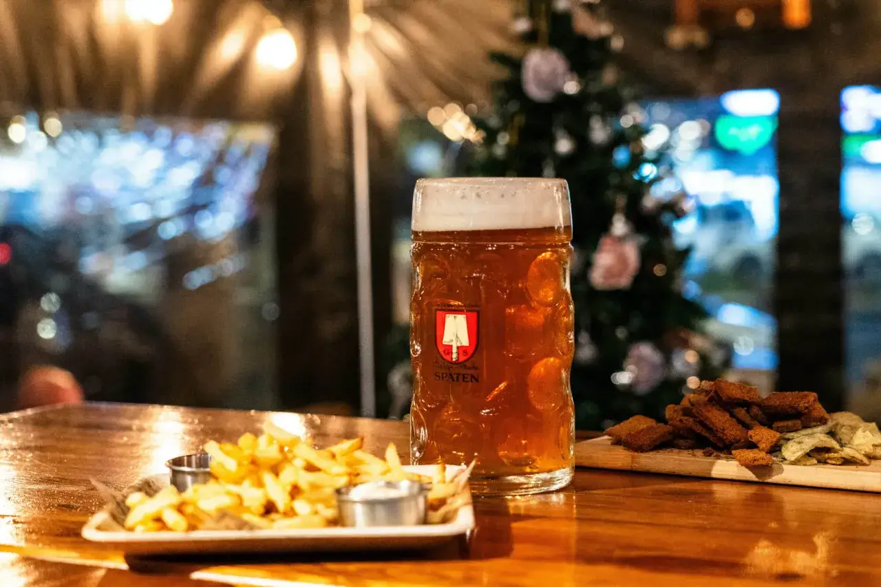 Une chope de bière ambrée, des frites et des croutons sur une table en bois, avec un sapin de Noël décoré en arrière-plan. Parfait pour une soirée bière chaude Noël.