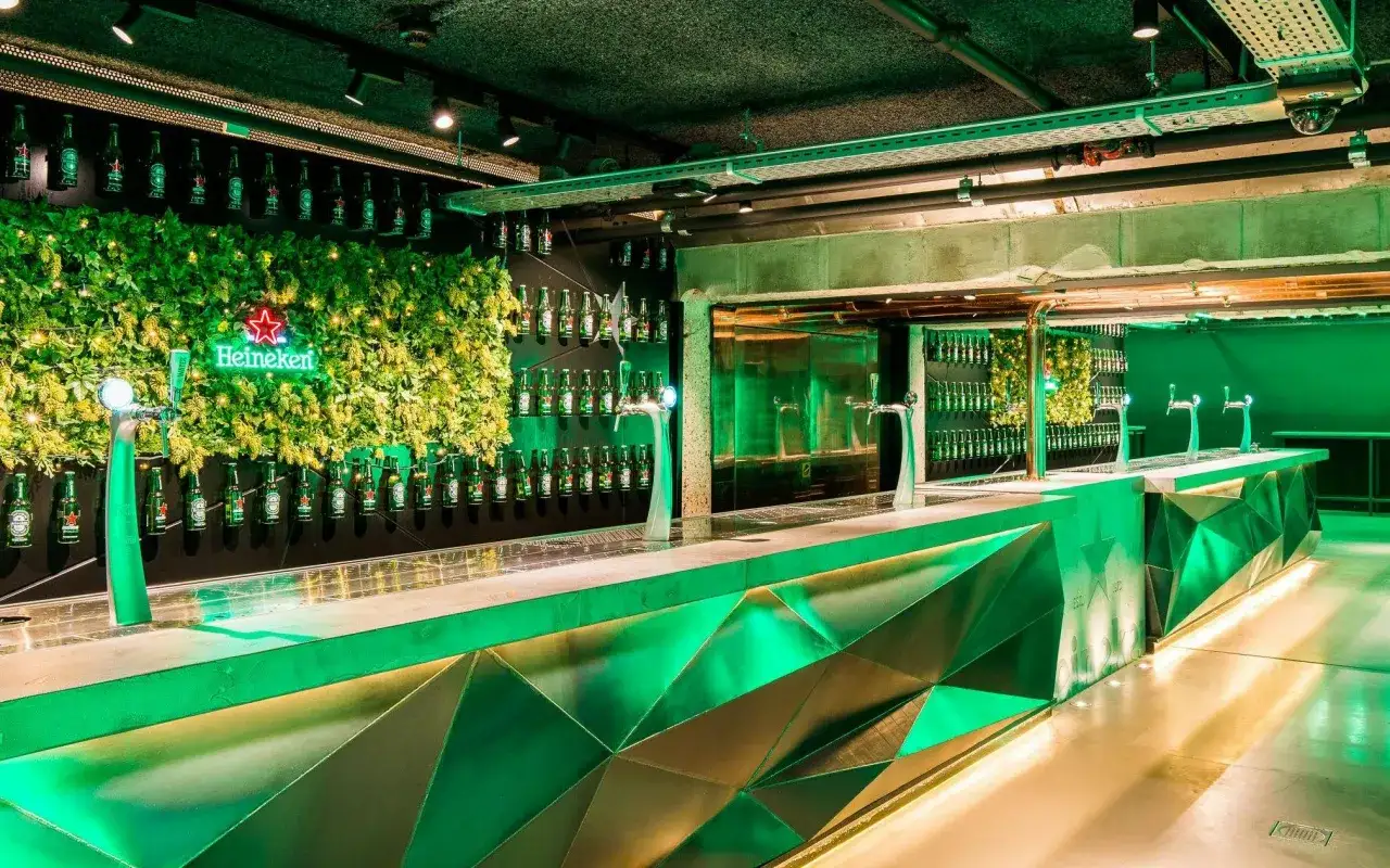 Bar moderne aux couleurs de Heineken, avec mur v&eacute;g&eacute;tal, n&eacute;on et bouteilles align&eacute;es. L'origine de la bi&egrave;re mise en avant.
