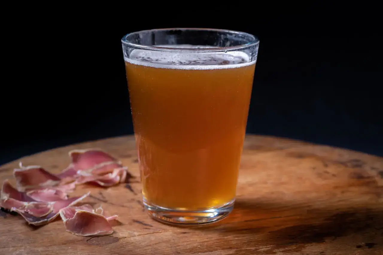 Verre de bière norvégienne ambrée, mousse blanche, sur une table en bois avec des tranches de viande séchée.